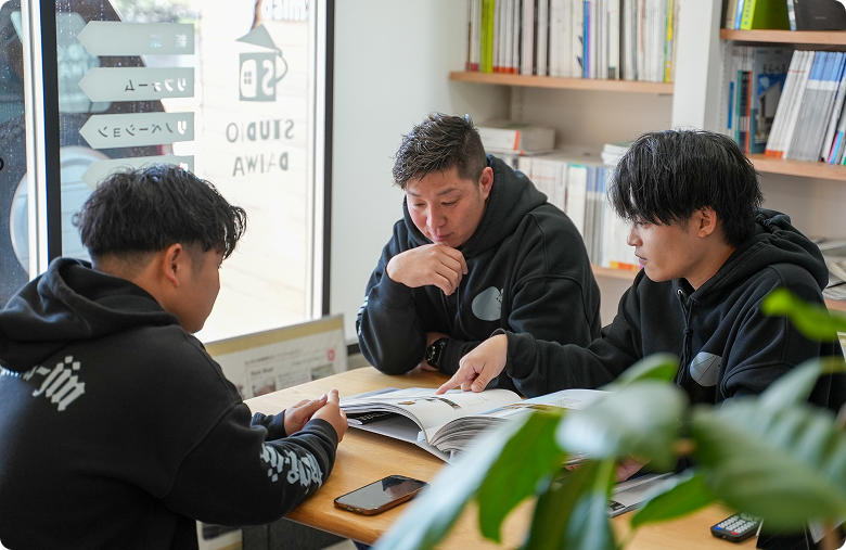 黒いパーカーを着た3人の男性が本棚のある明るいスペースで、資料を見ながら話し合っている。